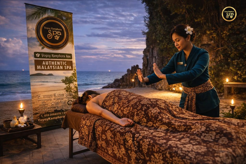 A professional promotional photo of a beachside massage at Yoyoy Kamphora Spa in Cherating. A female therapist in traditional blue attire and a batik sarong performs a treatment on a guest lying on a massage table draped in floral batik fabric. The scene is set on a sandy beach at dusk, illuminated by warm tiki torches with the ocean and a rocky cliff in the background. A tall promotional banner stands to the left.